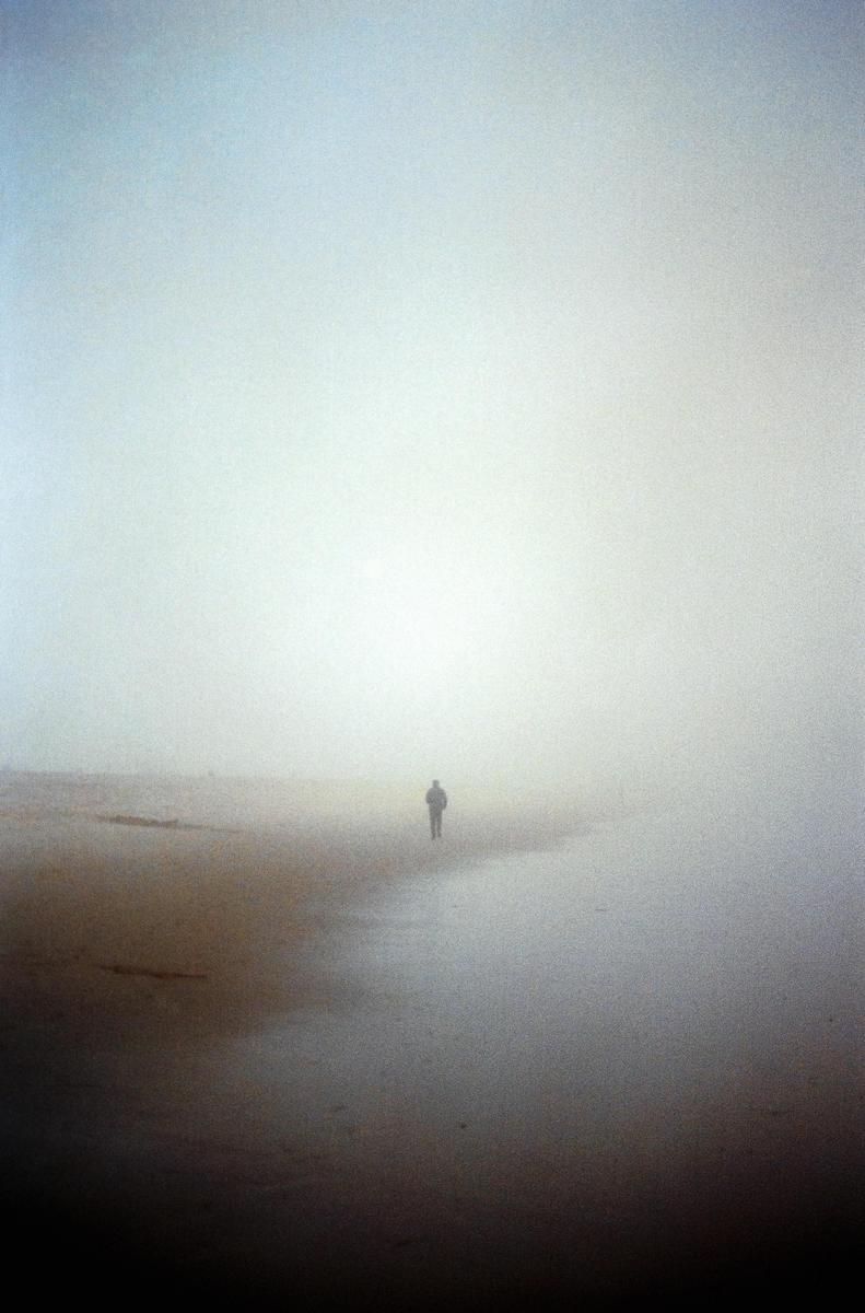 A lone man walks along a foggy beach.