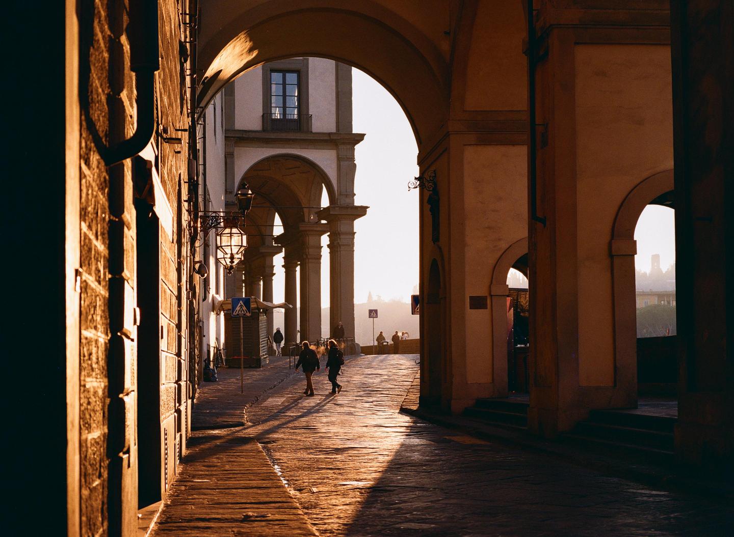 Two people walk on a sunlit cobblestone street under an archway in Florence.