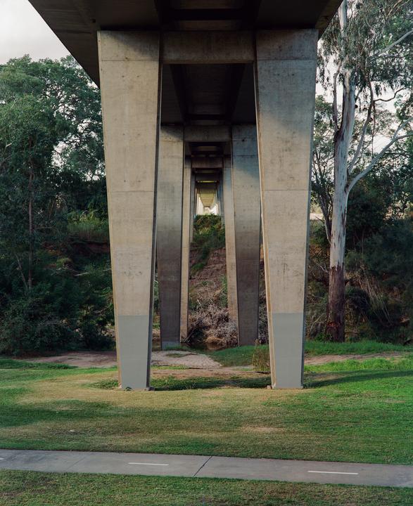A concrete bridge extends over a grassy area with trees and a small stream.