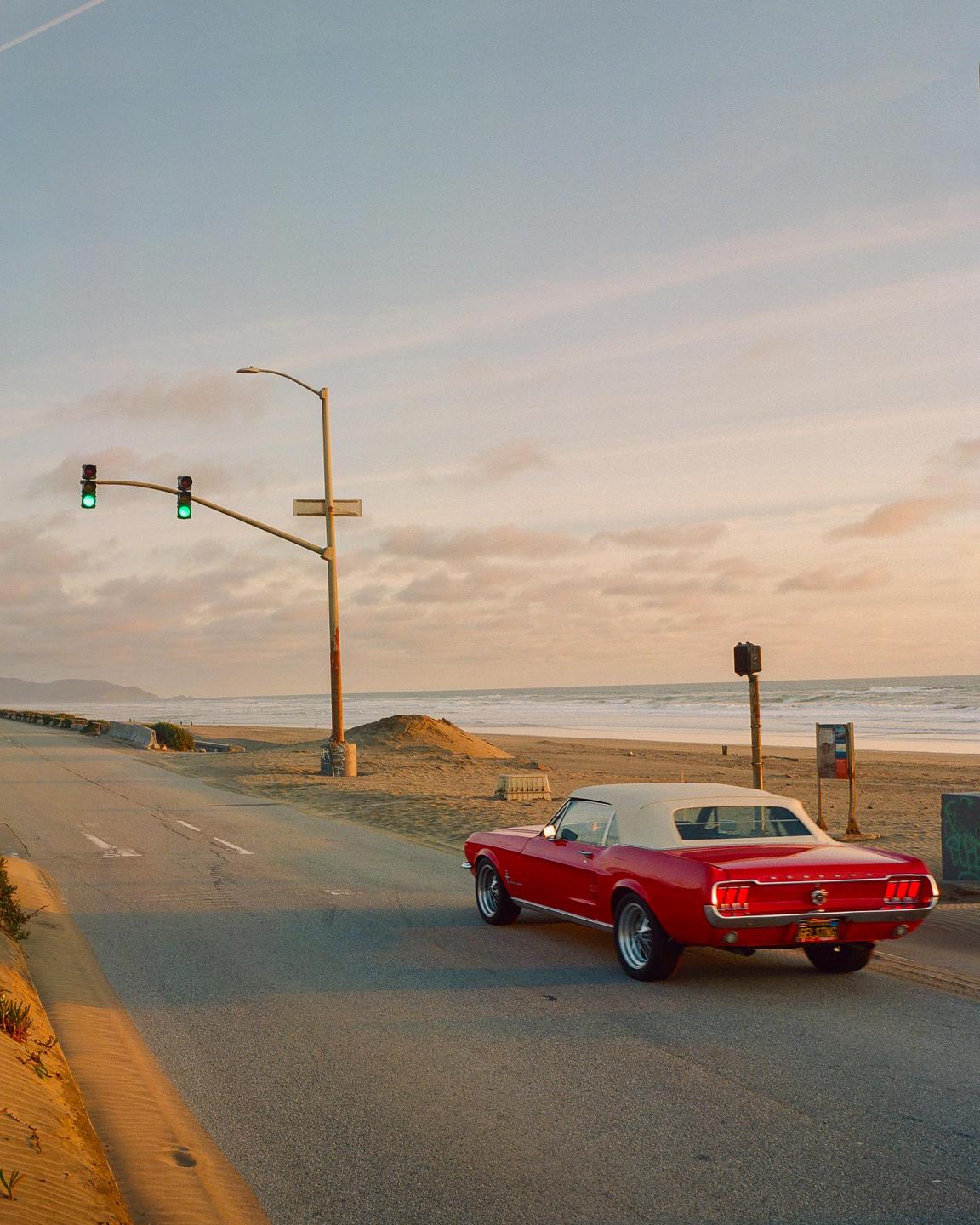 A red convertible car is parked on the side of a road next to a sandy beach at sunset.