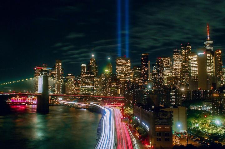 A city skyline at night features illuminated buildings, a bridge, and light trails from traffic on a highway.