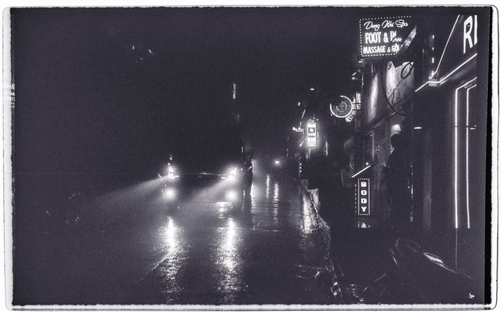 Headlights illuminate a wet street at night with neon signs on the right.