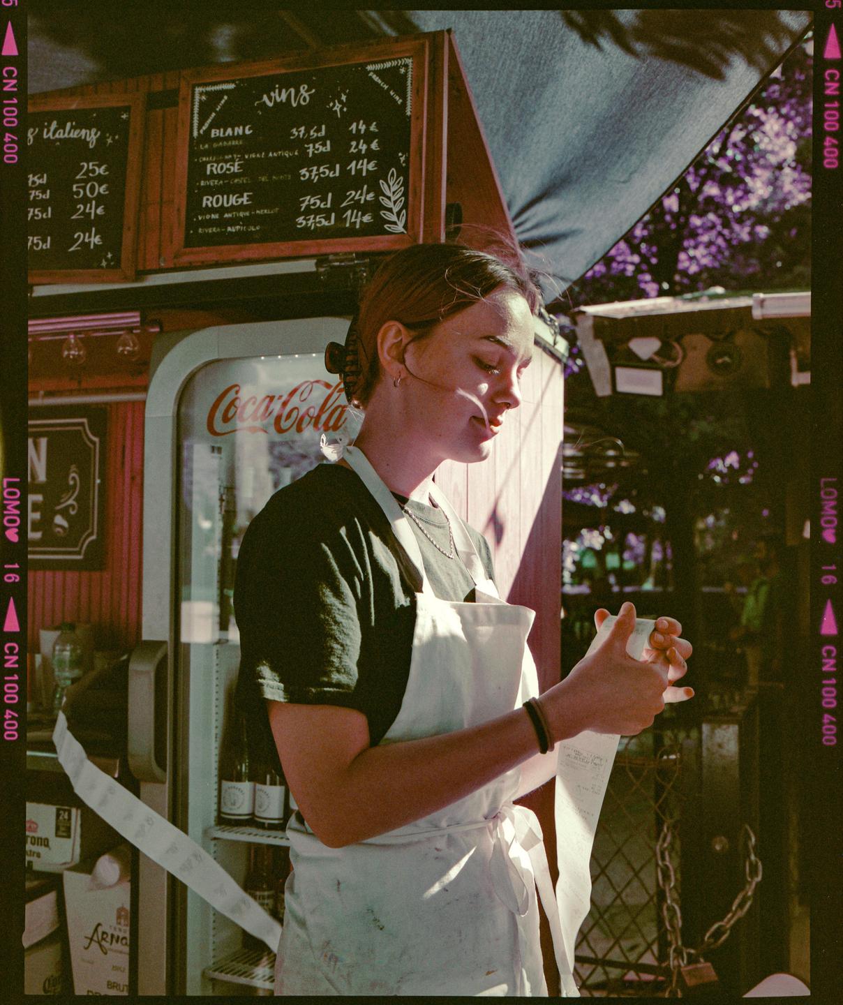 A woman in an apron stands next to a Coca-Cola refrigerator and a menu board, holding a long receipt.