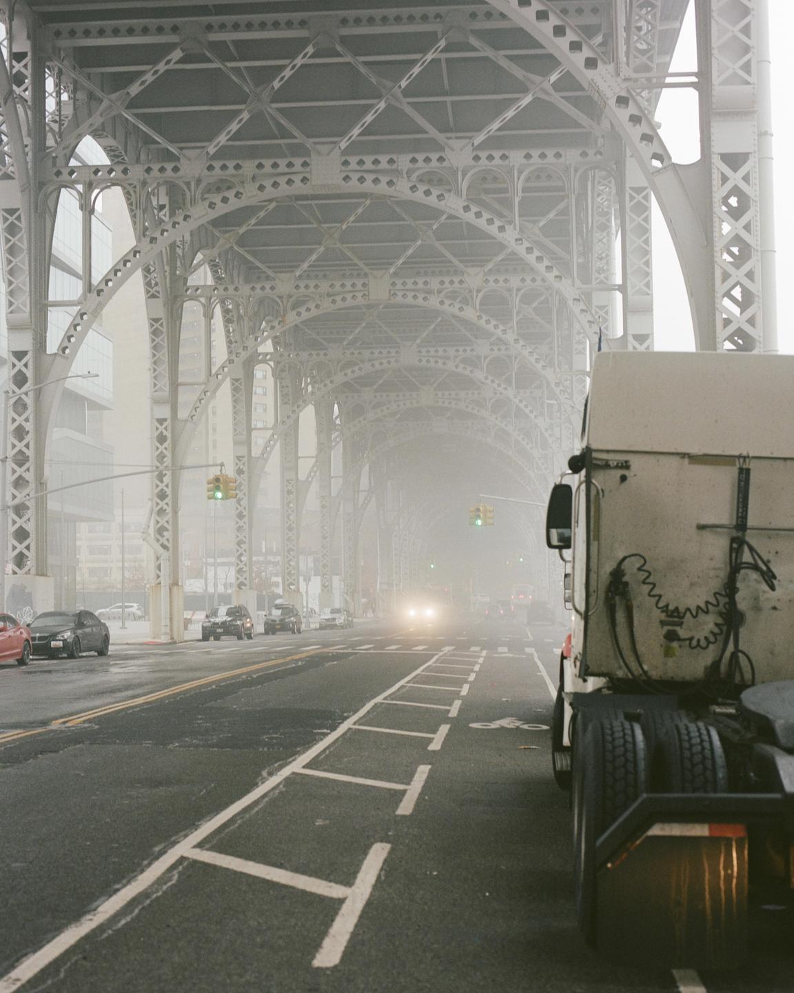 A truck is parked on the side of a road under a large metal bridge on a foggy day.