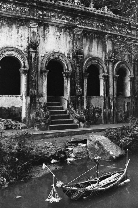 A small boat floats in a pond in front of an ornate, weathered building with arched doorways and a staircase.