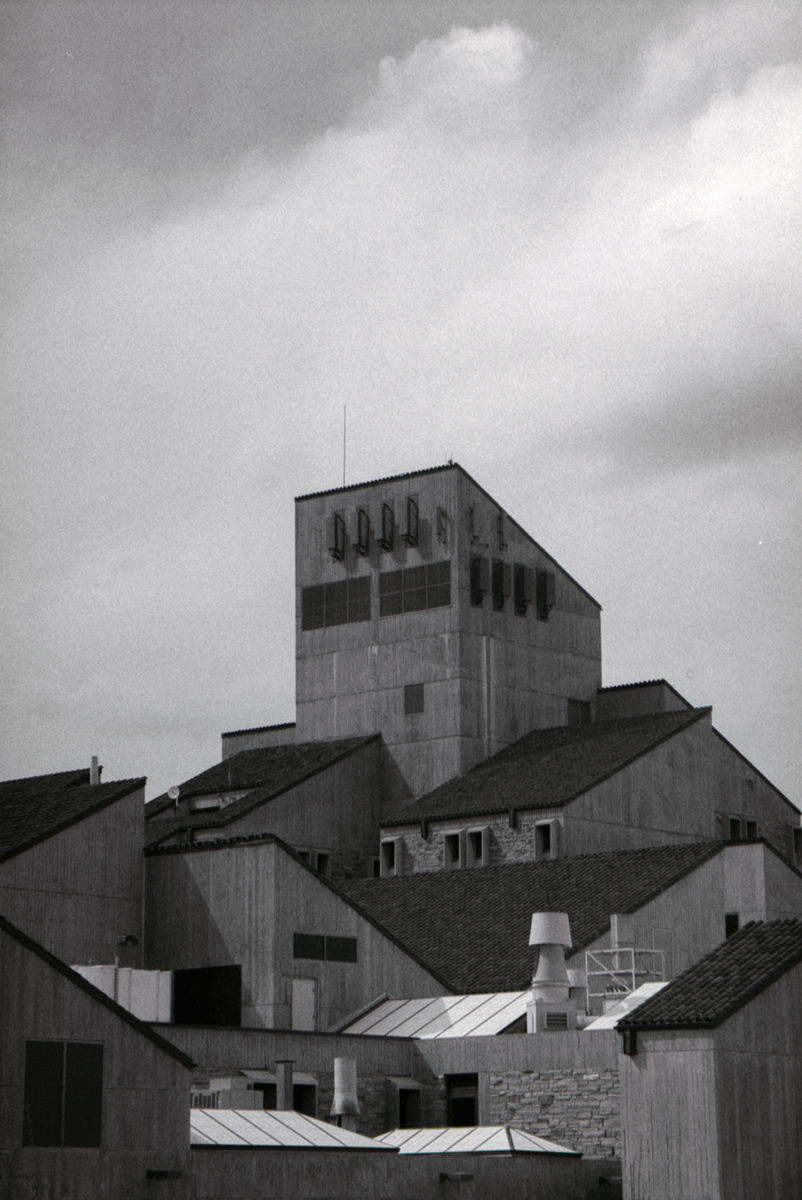 A tall concrete building with a slanted roof stands among other buildings with various rooflines under a cloudy sky.