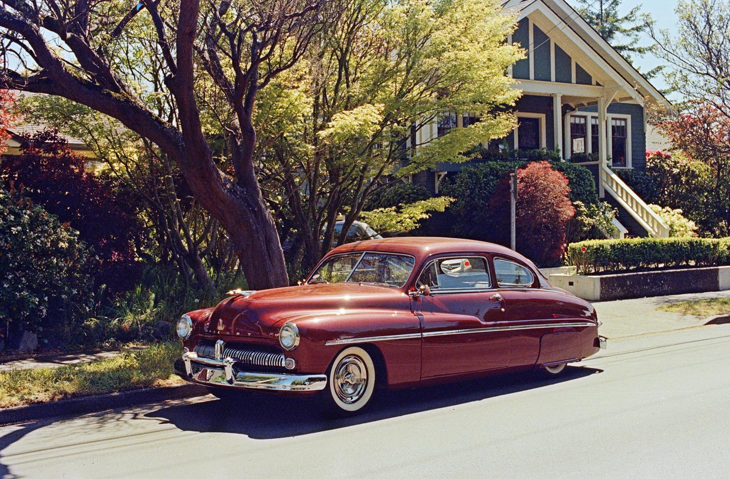 A shiny red vintage car is parked on a residential street in front of a house with a large tree.