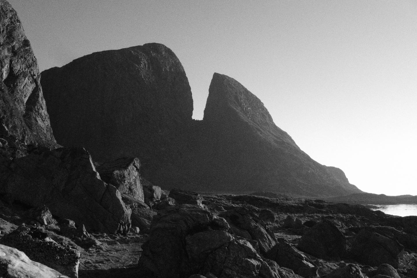 A black and white landscape shows a rocky foreground leading to a body of water with mountains in the background.