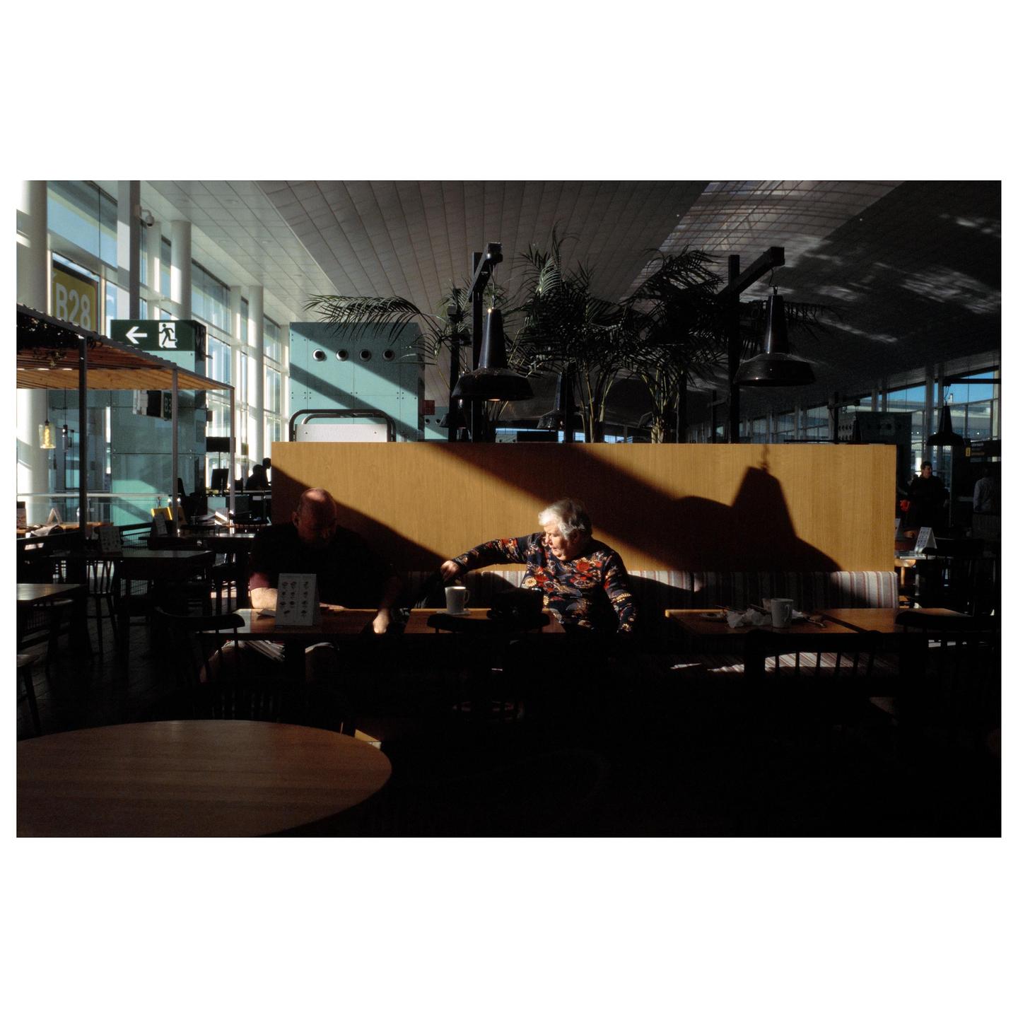 A man and a woman sit at a table in a cafe with strong shadows cast across the wall behind them.