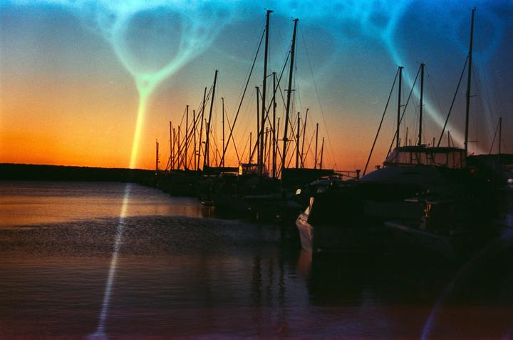 A marina full of sailboats and yachts is silhouetted against a vibrant sunset with a bright light streak across the sky and water.