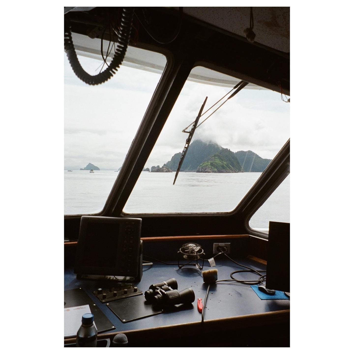 A view from inside a boat's cockpit shows a mountainous island in the distance under a cloudy sky.