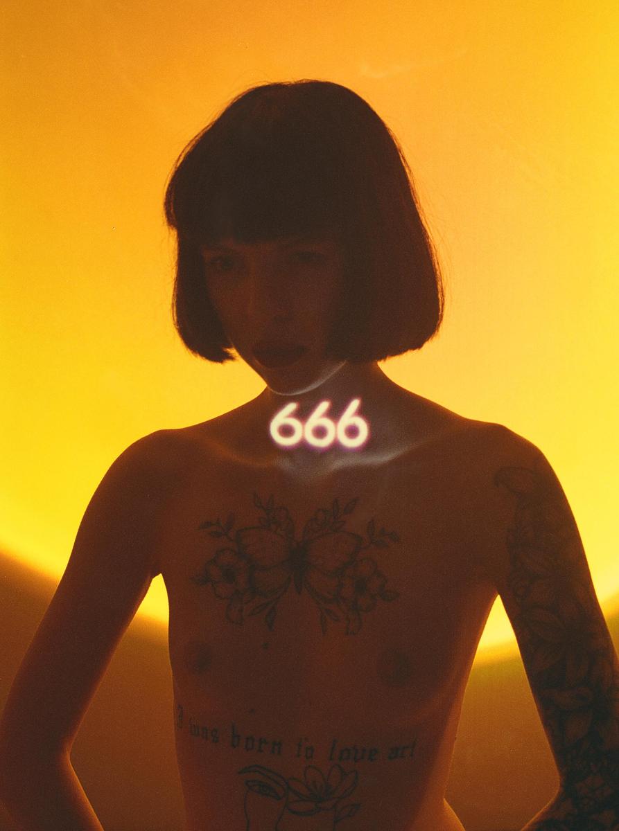 A woman with short hair and tattoos on her chest and arms has the number 666 projected onto her neck.