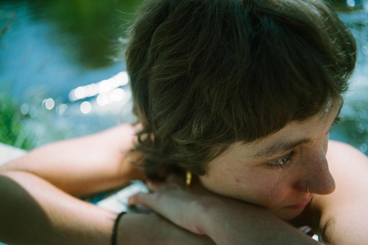 A woman with short hair and a nose ring rests her head on her arms, looking towards a body of water with bright reflections.