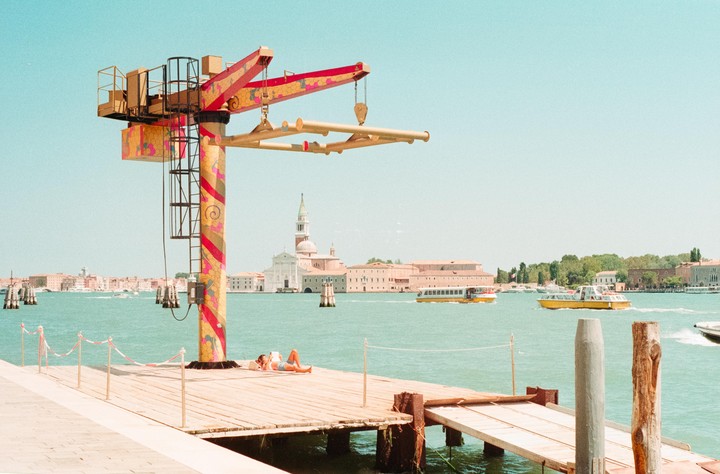 A person sunbathes on a wooden pier next to a colorful crane, with boats on the water and a city in the background.