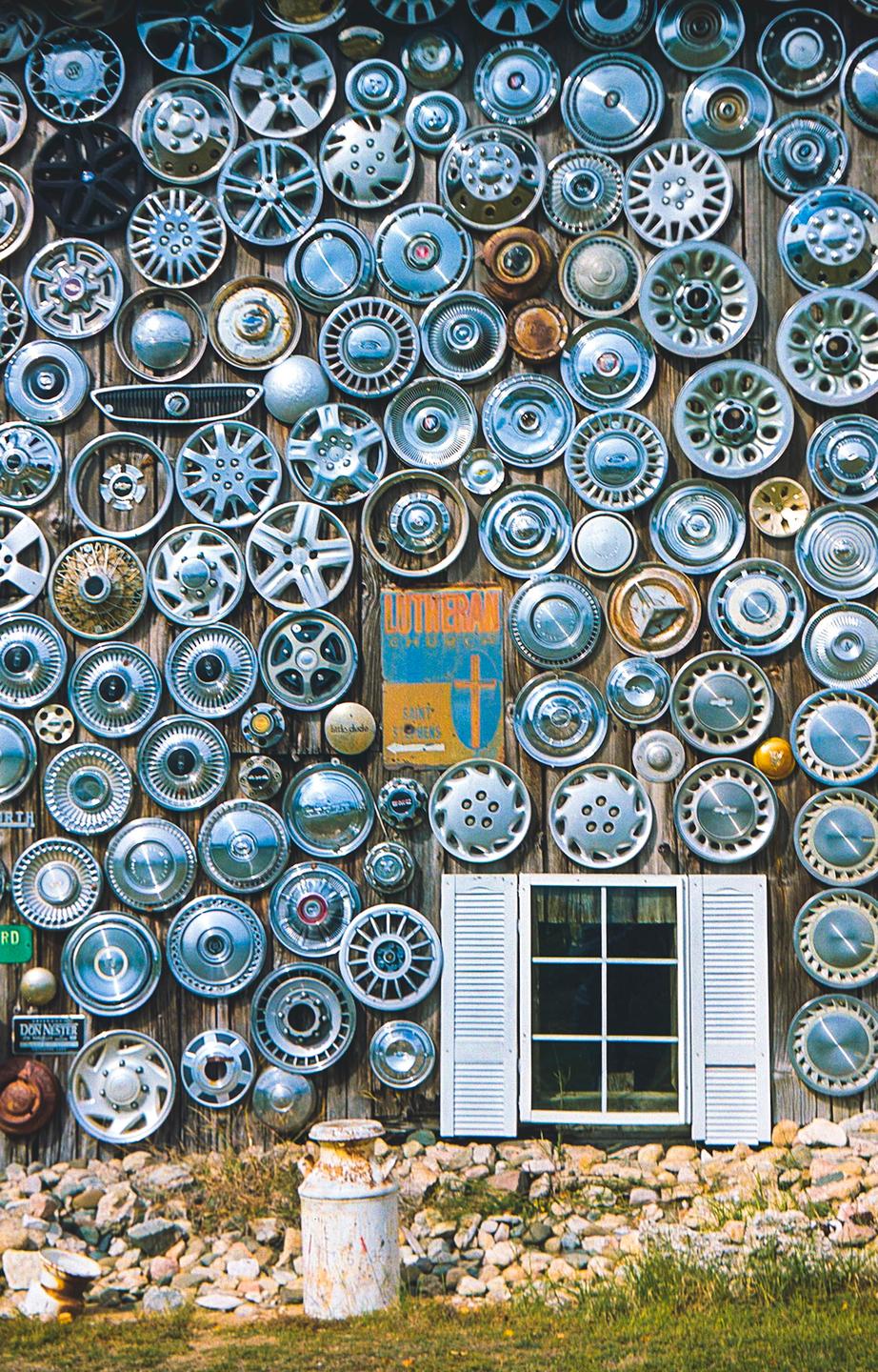 A wooden wall is covered with many hubcaps and a white-shuttered window, with a milk can and rocks at its base.