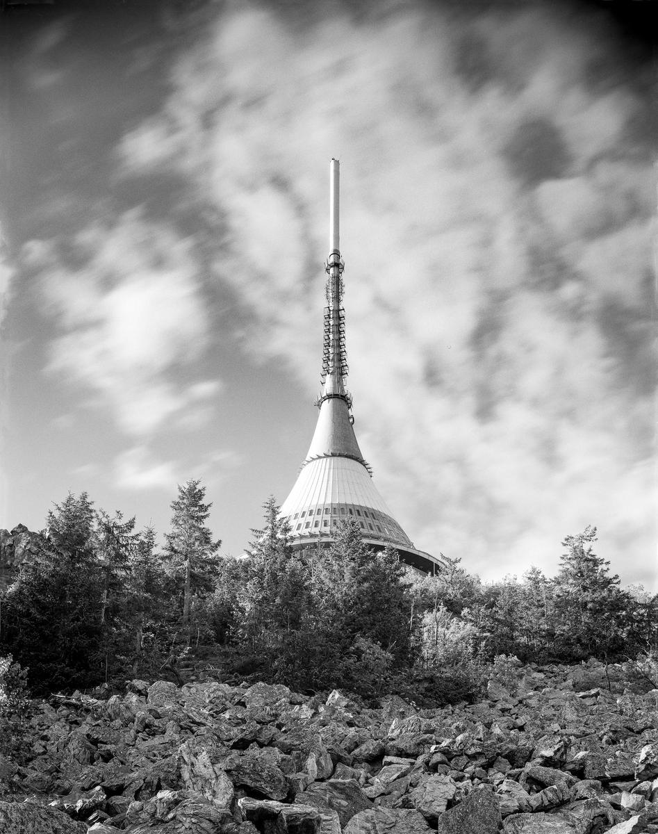 A tall, conical tower with a spire rises above a rocky, forested landscape under a cloudy sky.