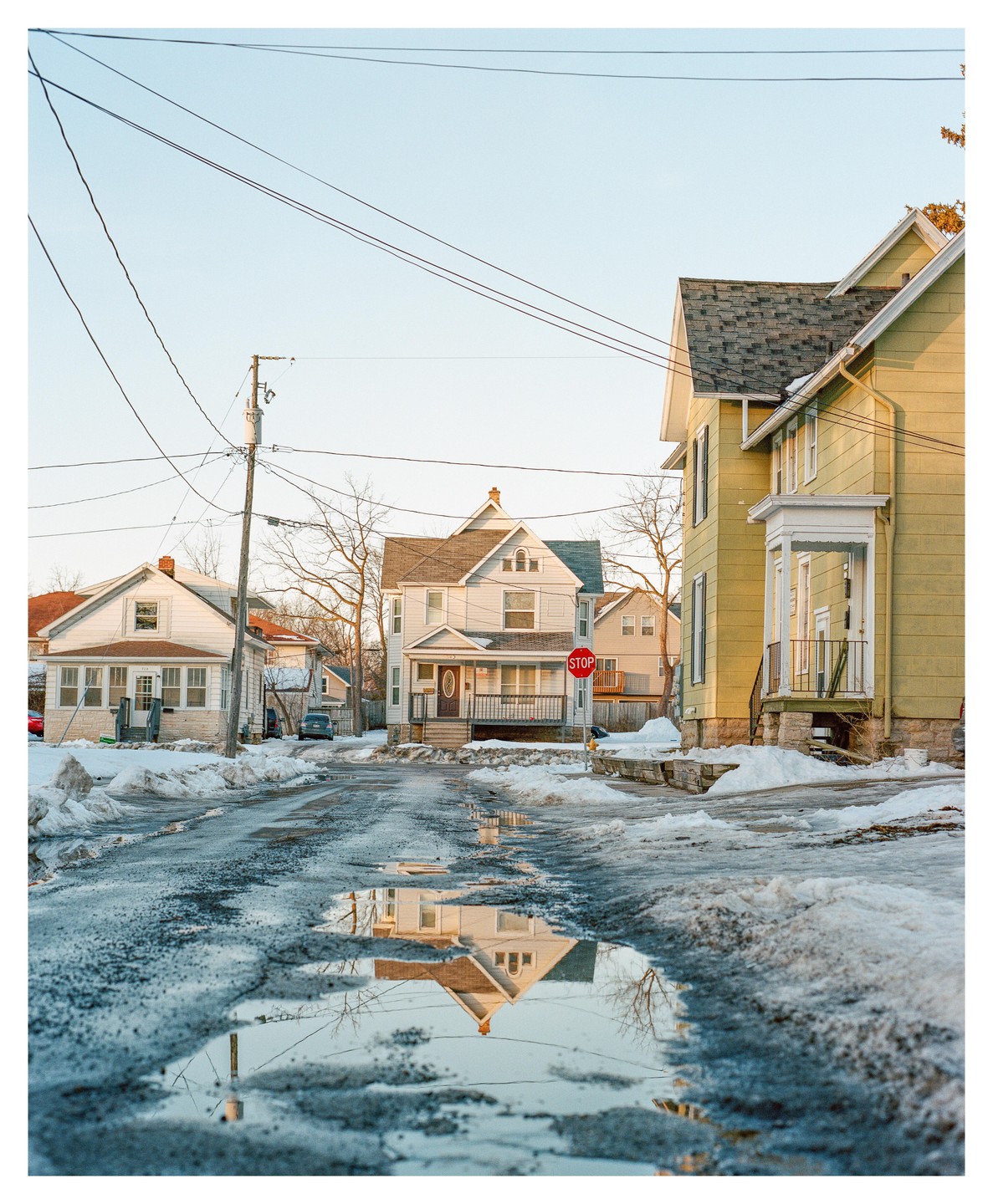A street with houses and power lines is covered in snow and puddles, reflecting a house and the sky.