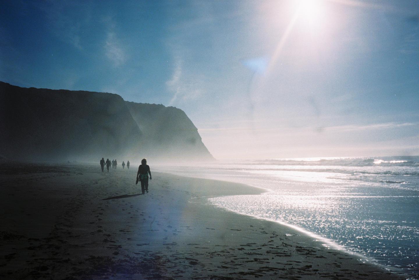 A group of people walk along a beach with cliffs in the background and bright sunlight reflecting on the water.