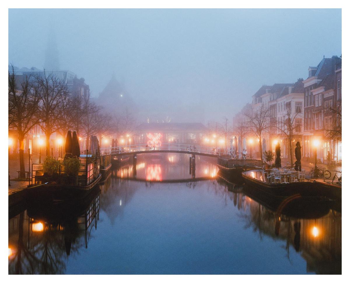 A canal with boats and bridges is surrounded by buildings and trees, with lights reflecting on the water through a thick fog.