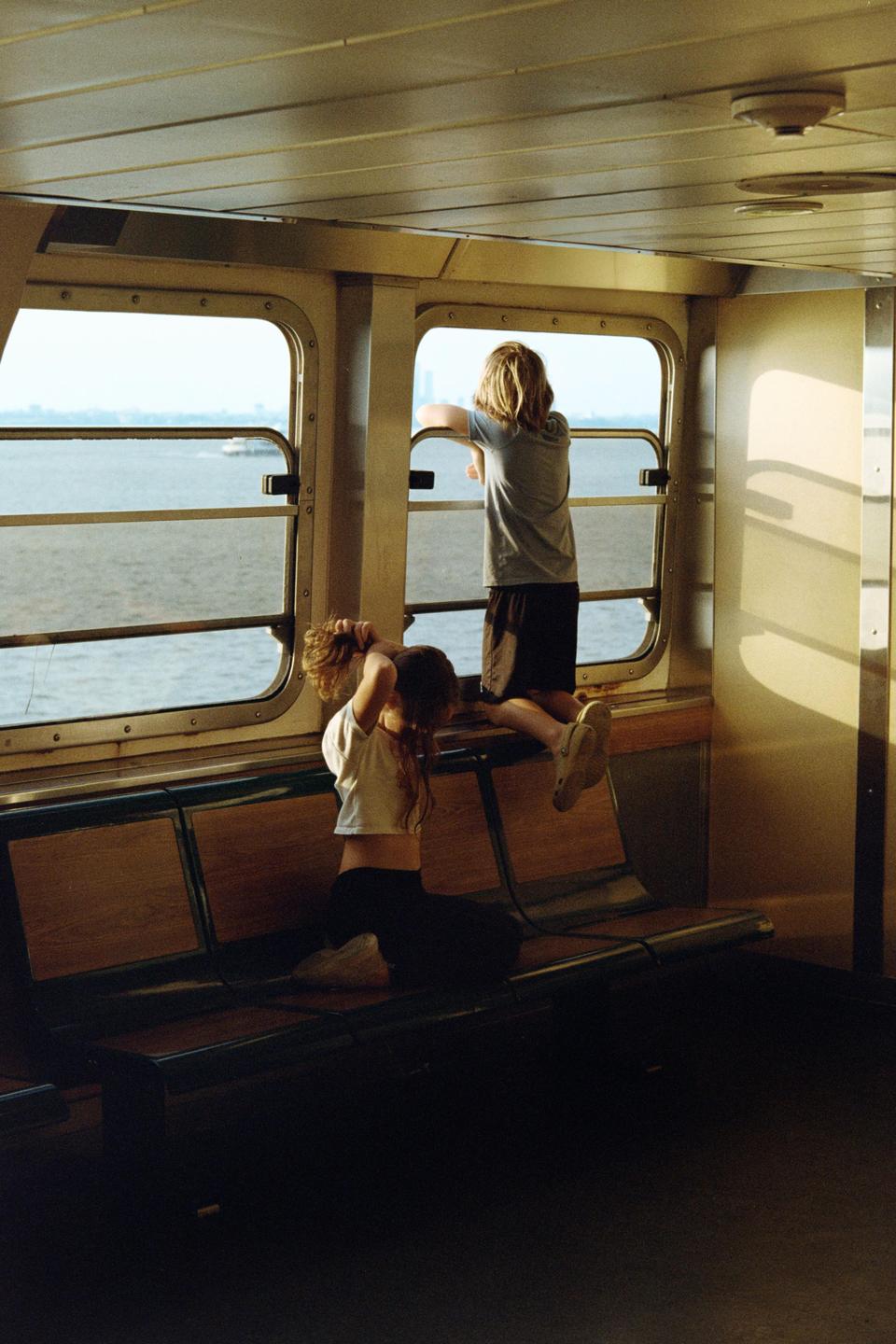 Two children are inside a boat, one looking out a window at the water and the other sitting on a bench.