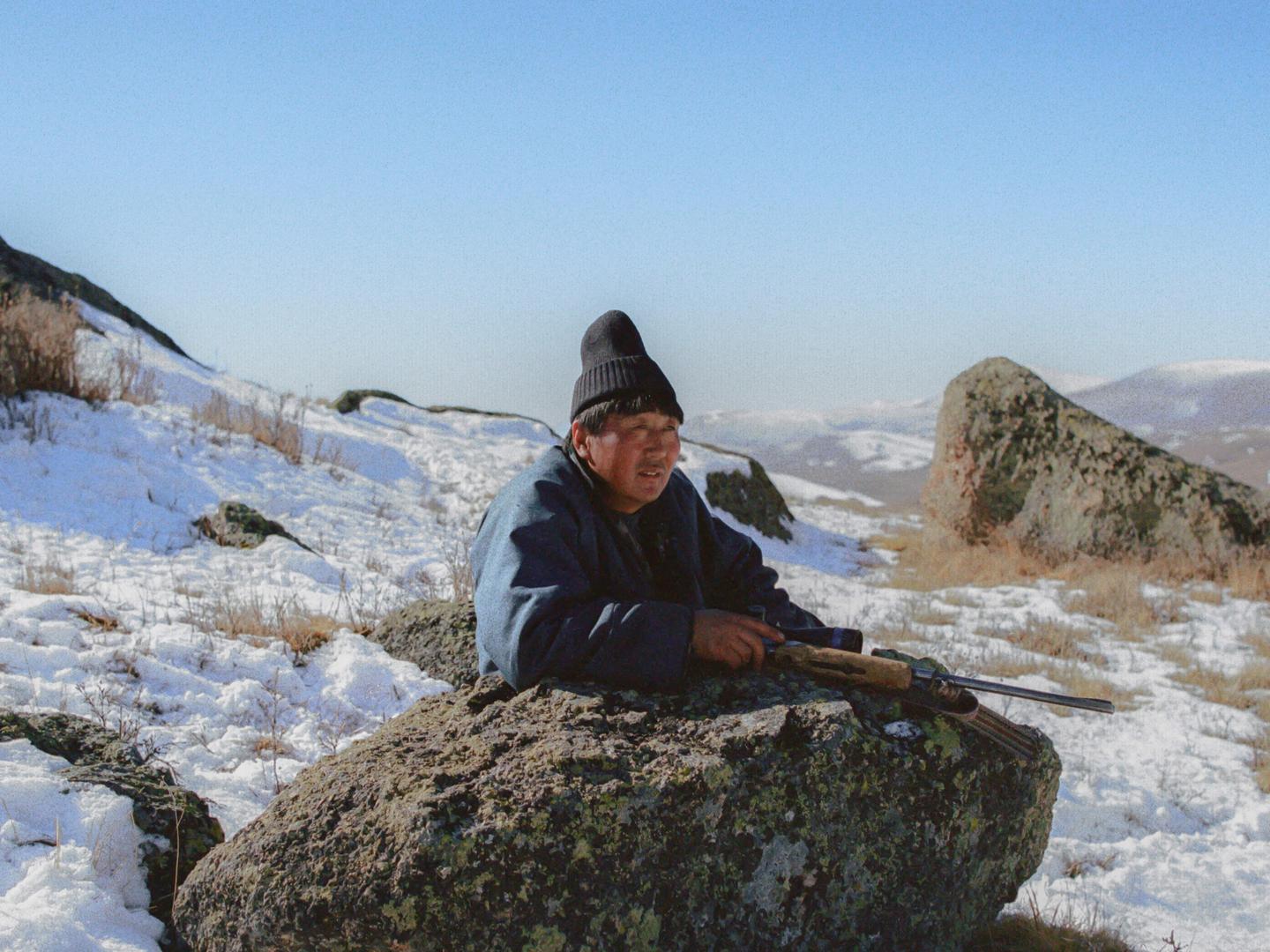 A man in a dark jacket and hat rests on a large rock with a rifle in a snowy, mountainous landscape.