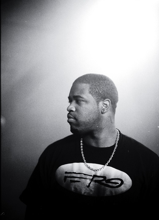 A man with a chain necklace and a t-shirt with a logo looks to the left.