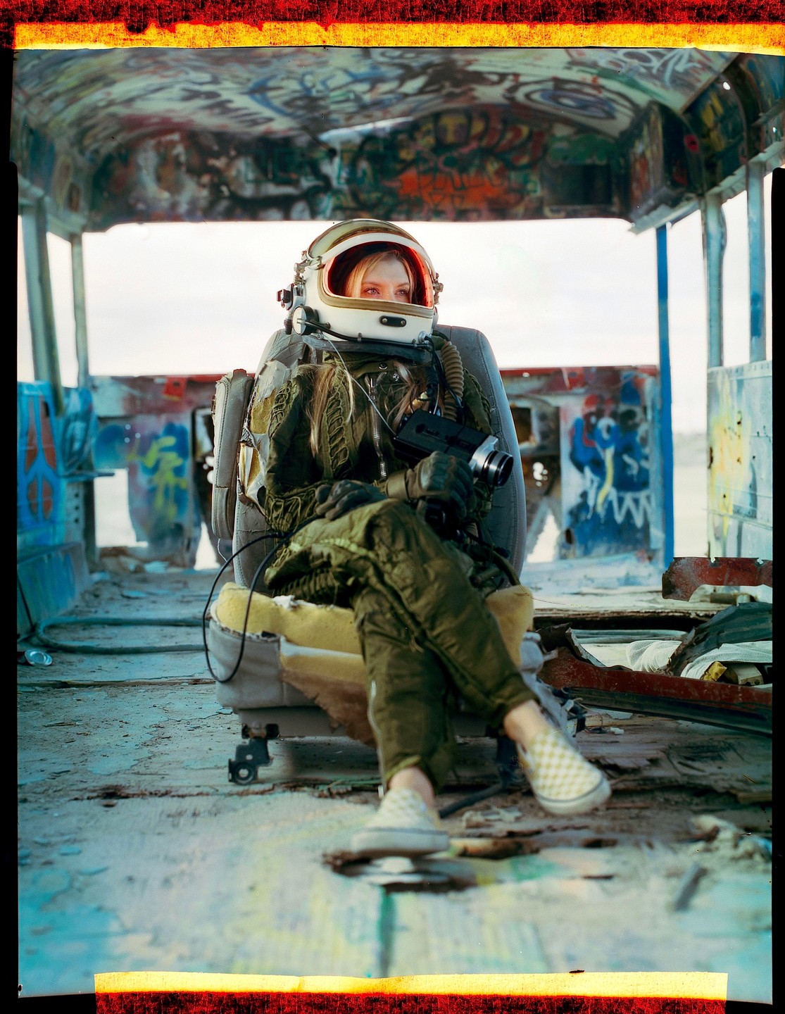 A woman in an astronaut suit sits in a derelict, graffiti-covered vehicle.