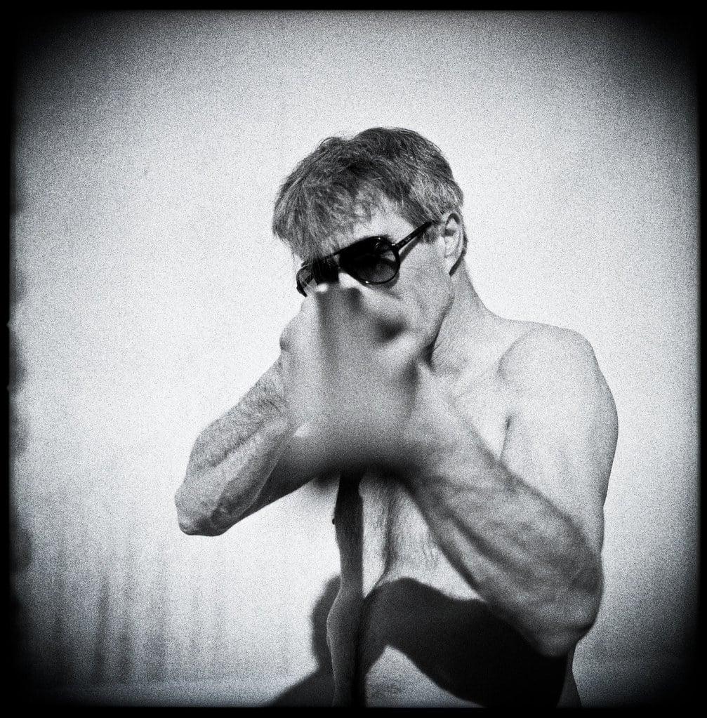 A shirtless man wearing sunglasses holds his hand up to his face in a black and white self-portrait.