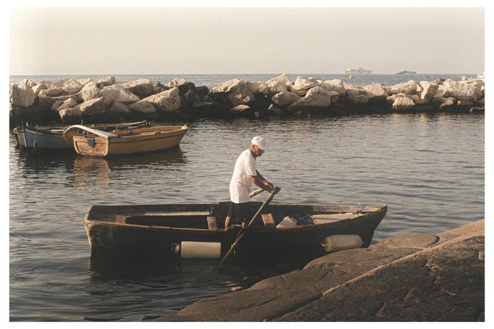 A man in a small boat rows near a rocky breakwater with other boats in the water.