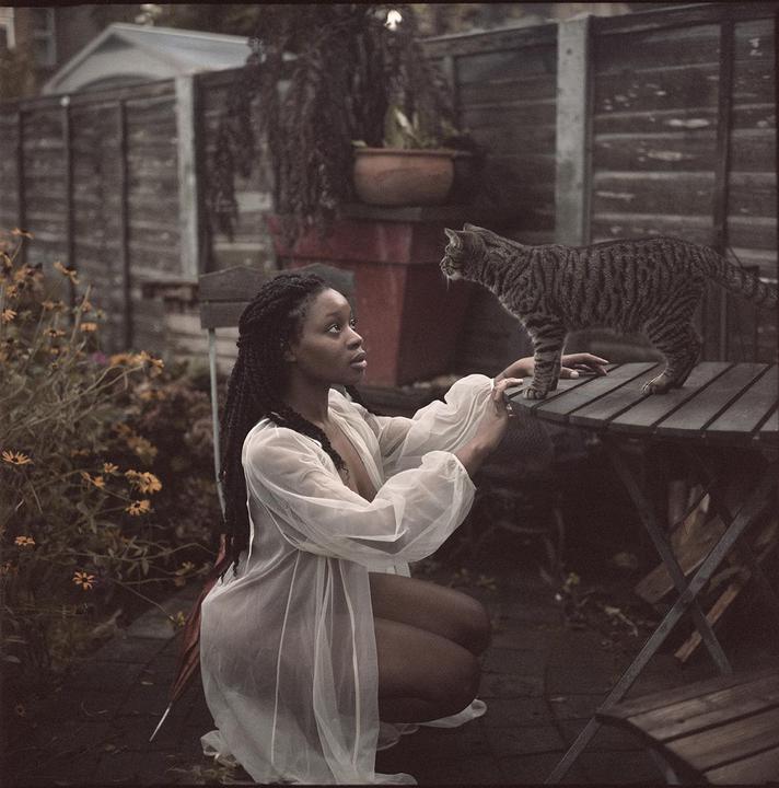 A woman in a sheer white robe crouches in a garden, looking up at a tabby cat standing on a wooden table.