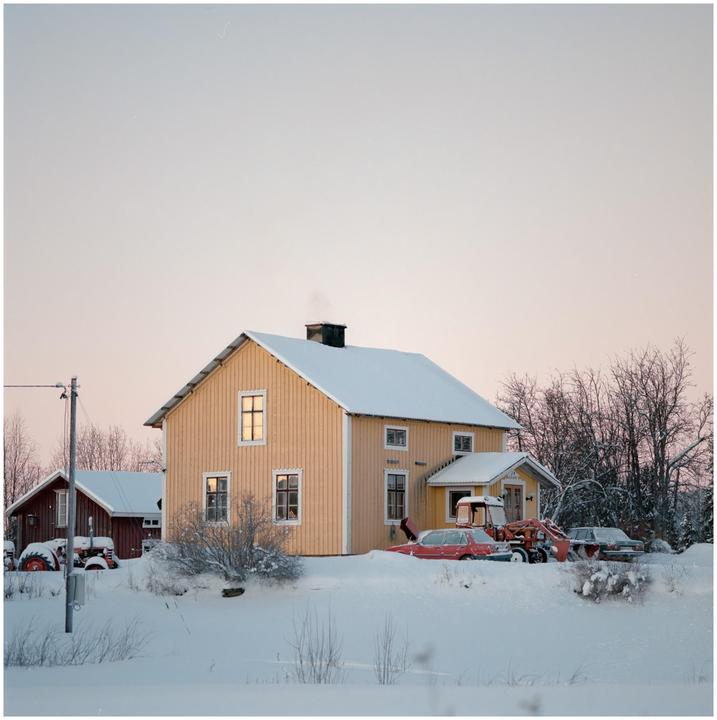 A yellow house with a snow-covered roof stands in a snowy landscape with trees and vehicles.