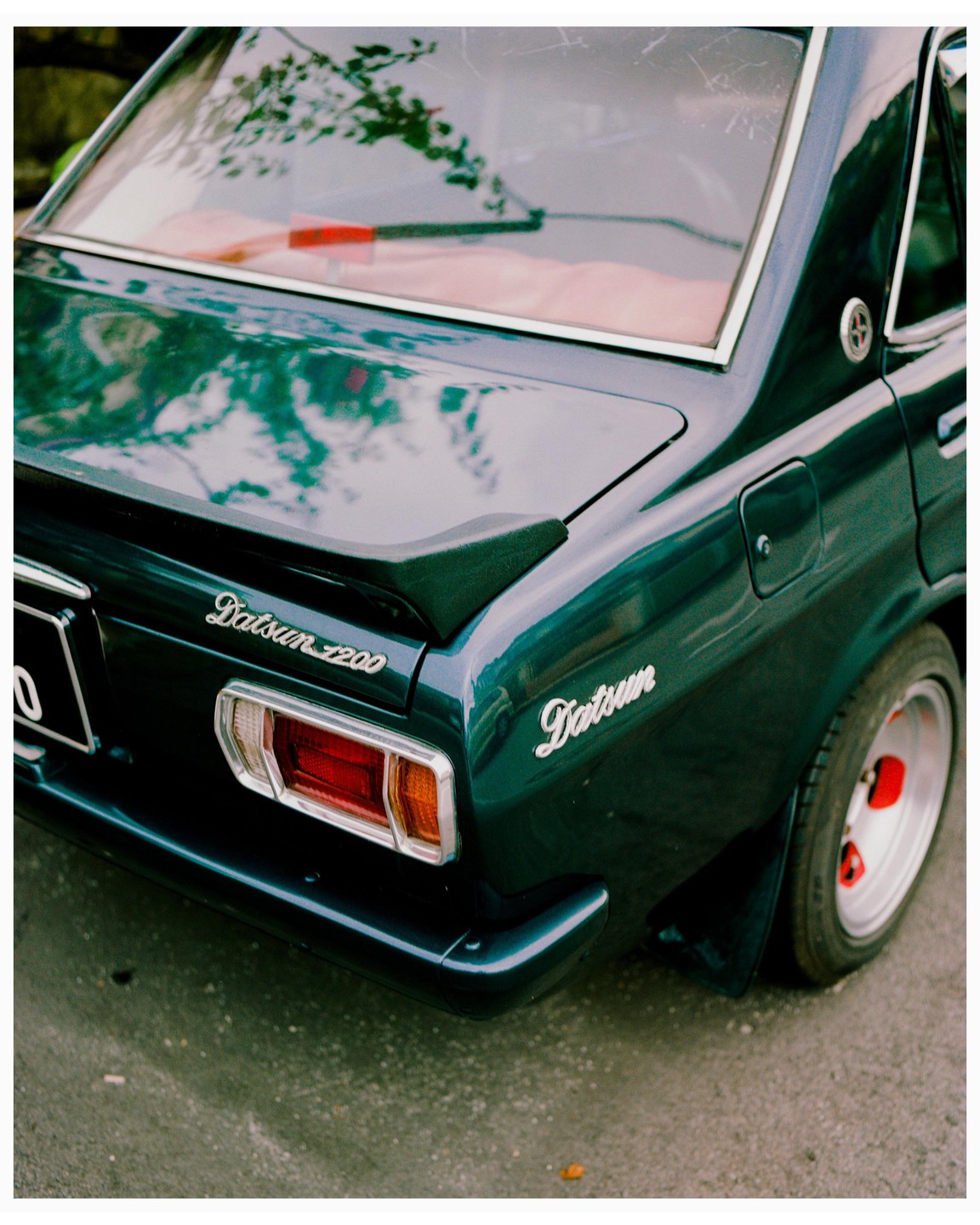 The rear of a dark green Datsun 1200 is parked on asphalt.
