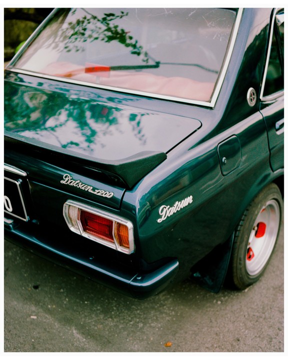 The rear of a dark green Datsun 1200 is parked on asphalt.