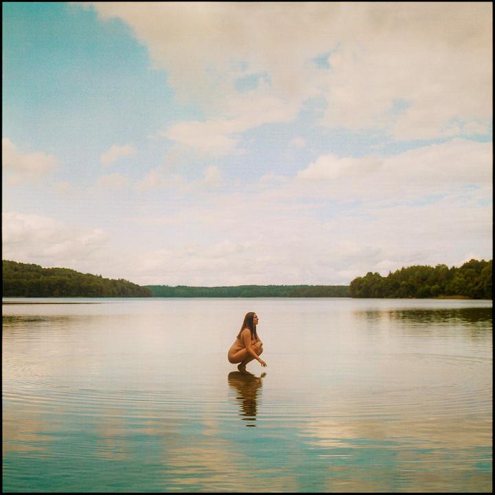 A woman crouches in a lake with trees on the far bank under a cloudy sky.