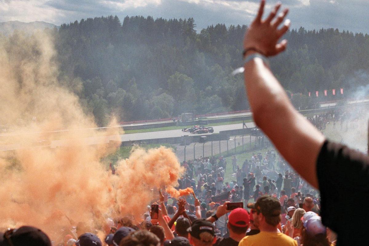 A crowd of spectators watches a car race from a hillside, with orange smoke rising from the crowd.