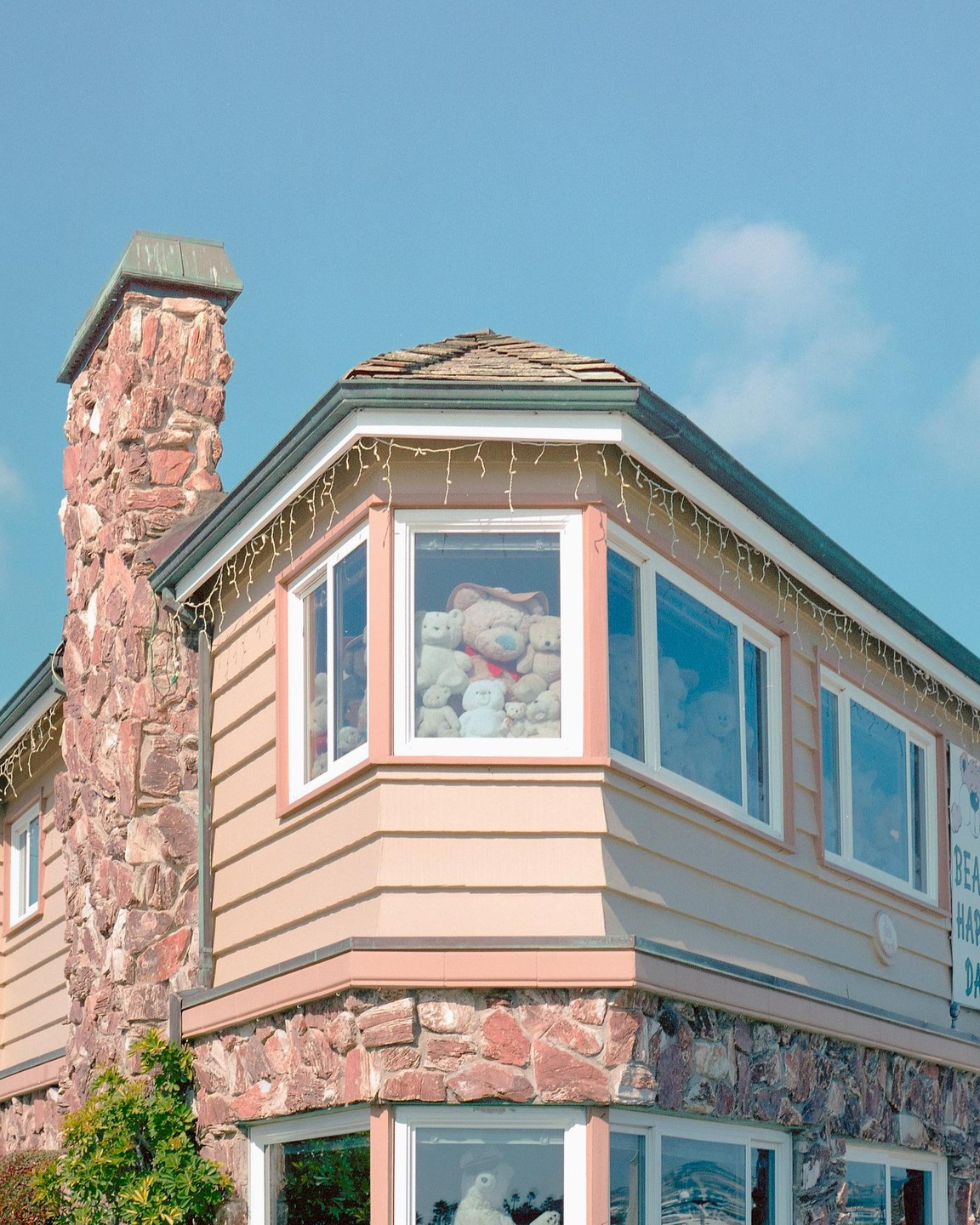 A house with a stone chimney and many teddy bears visible through the windows is shown under a clear blue sky.