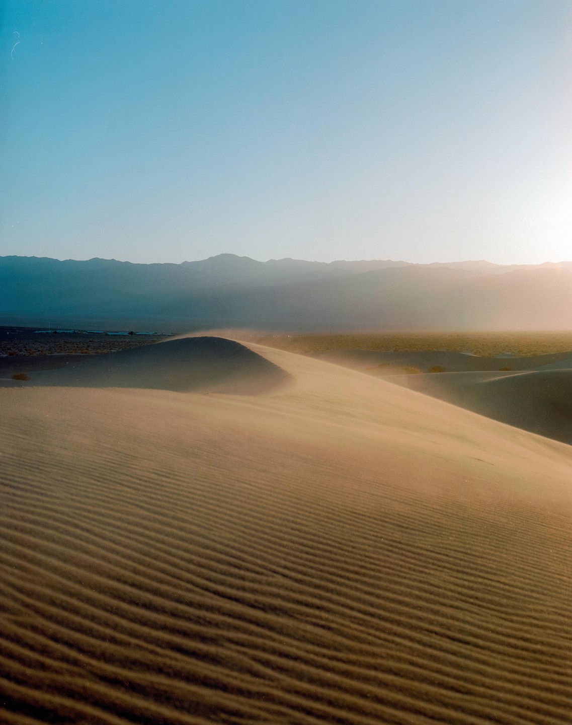 Wind blows sand across a desert dune with mountains in the distance under a clear sky.