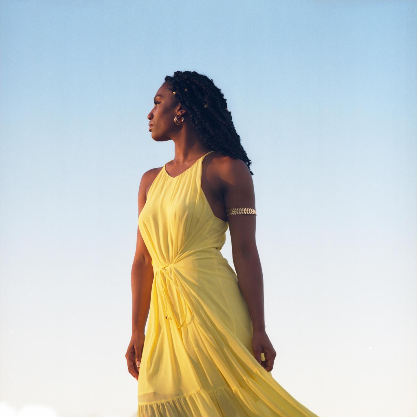 A woman in a yellow dress and an arm cuff looks to the side against a clear sky.