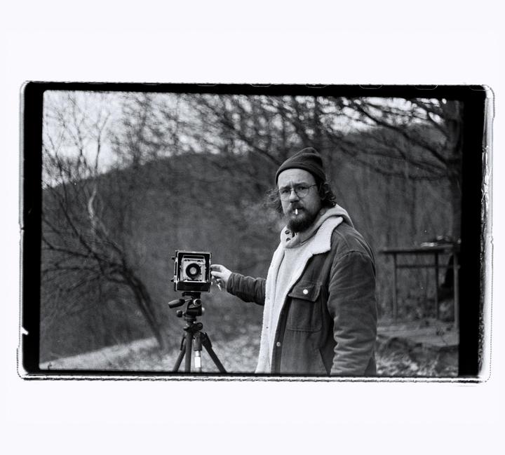 A man with a cigarette in his mouth stands next to a camera on a tripod in a wooded area.