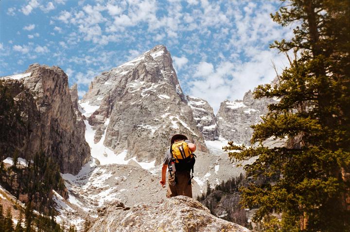 A man with a yellow backpack stands on a rock, looking up at snow-capped mountains under a blue sky with clouds.