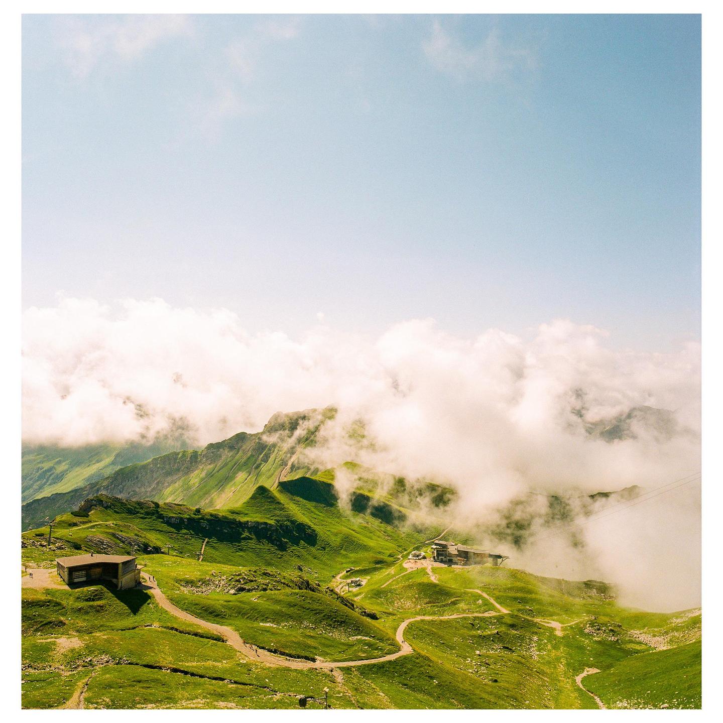 Clouds roll over green mountains with buildings and winding paths visible below.