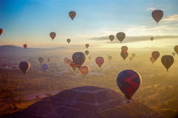 Hot air balloons float above a pyramid and a hazy landscape at sunrise.