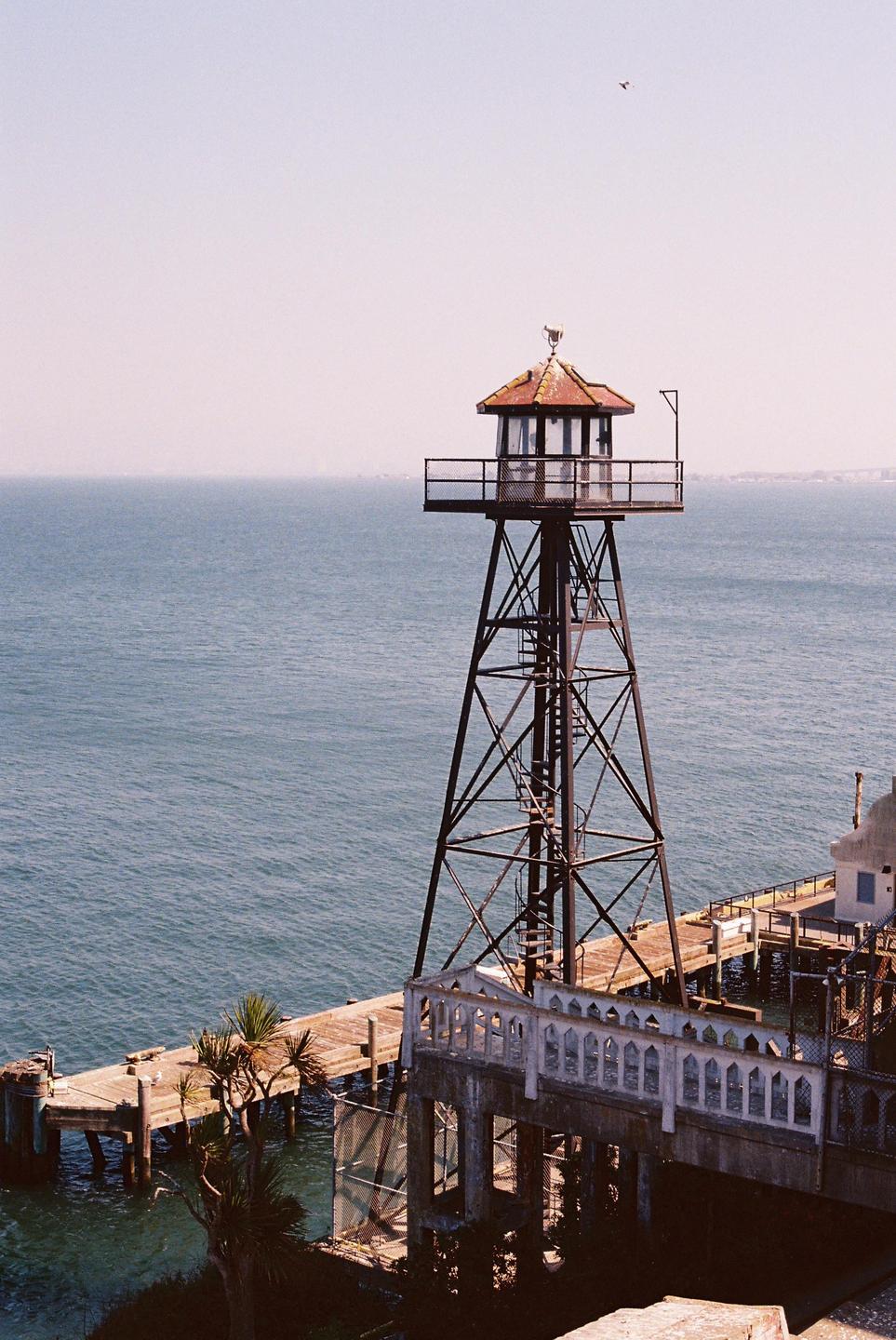 A tall metal tower with a small building on top stands next to a wooden pier over the ocean.