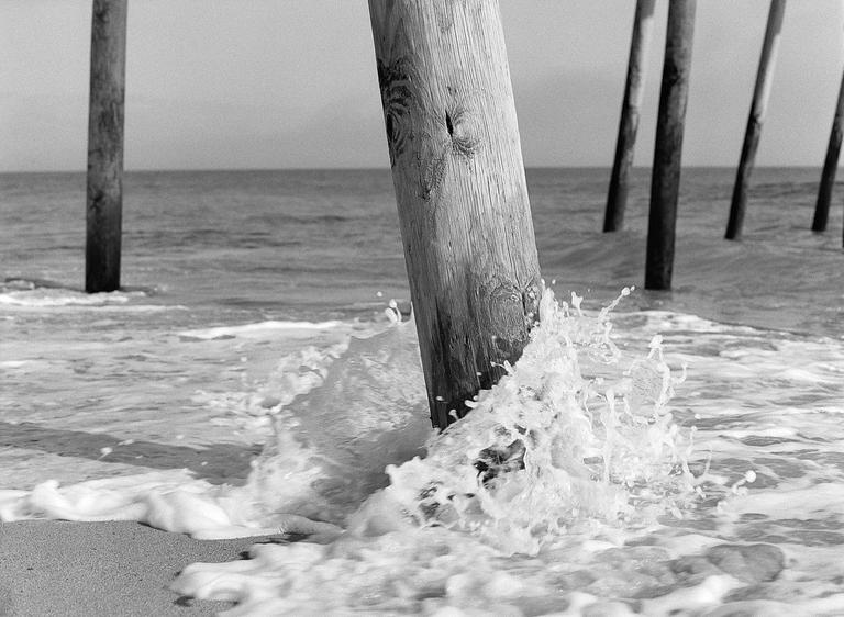 A wooden pier piling is splashed by a wave on a sandy beach.