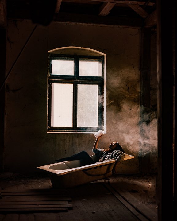 A woman reclines in a bathtub in a derelict room, holding a cloud of smoke above her.