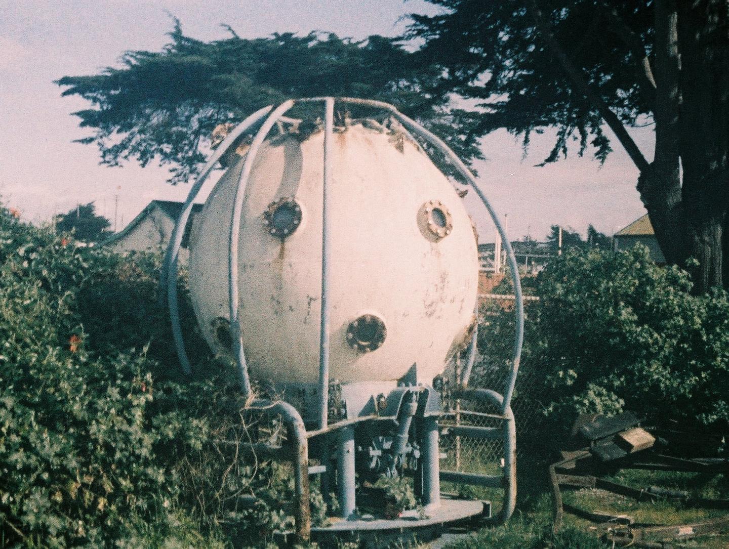 A large, spherical, white diving bell with portholes is surrounded by green bushes and trees.