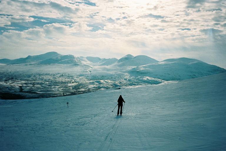 A person skis across a snowy landscape with mountains in the background under a cloudy sky.