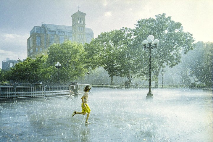 A child runs through a sudden downpour in a city square, with a large building and trees in the background.