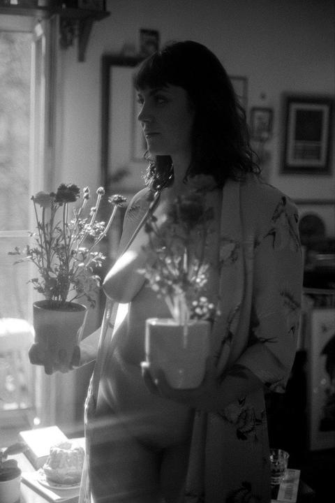 A woman in a robe holds two potted plants in a sunlit room.
