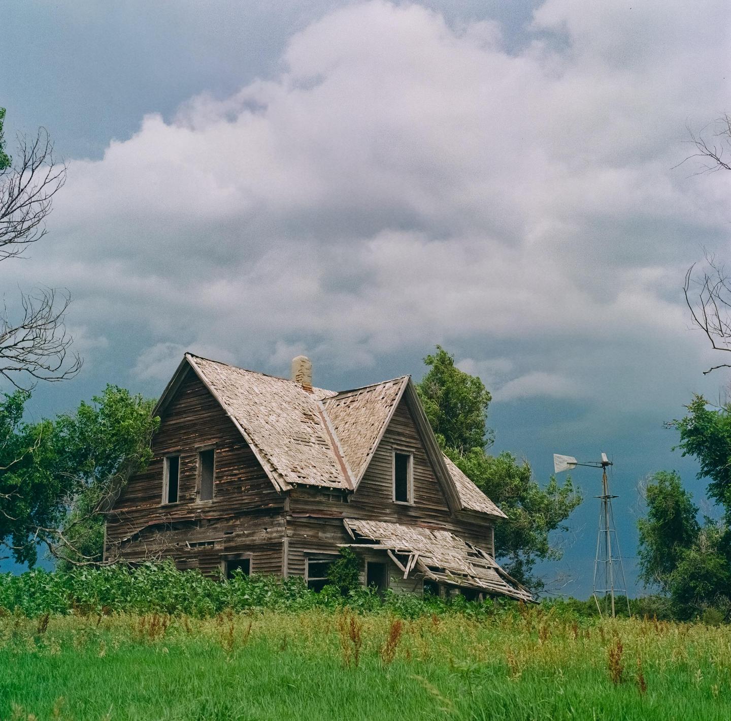 An abandoned wooden house with a damaged roof stands in a field under a cloudy sky, with a windmill visible in the distance.